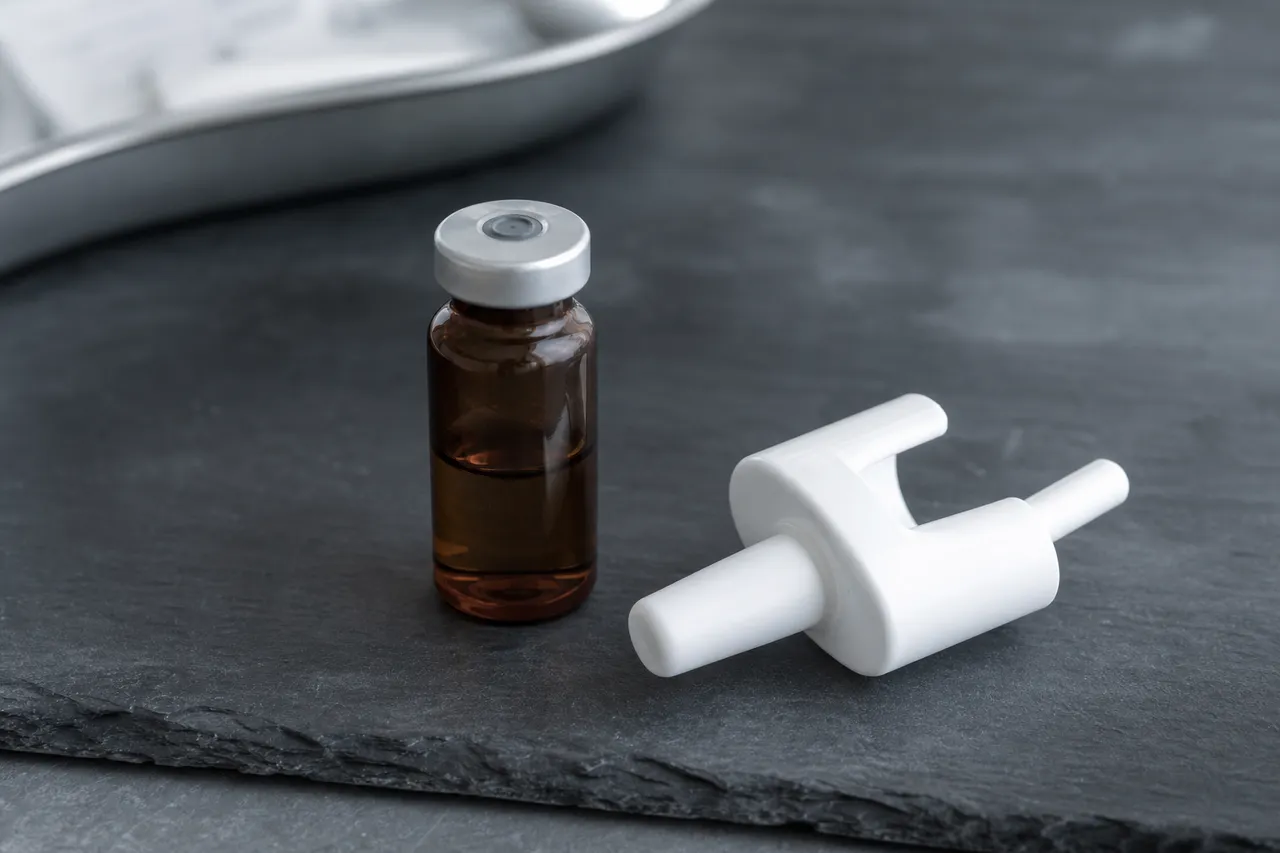 Editorial still life of a small amber pharmaceutical glass vial with a rubber stopper on a dark slate surface, next to a white naloxone nasal-spray device