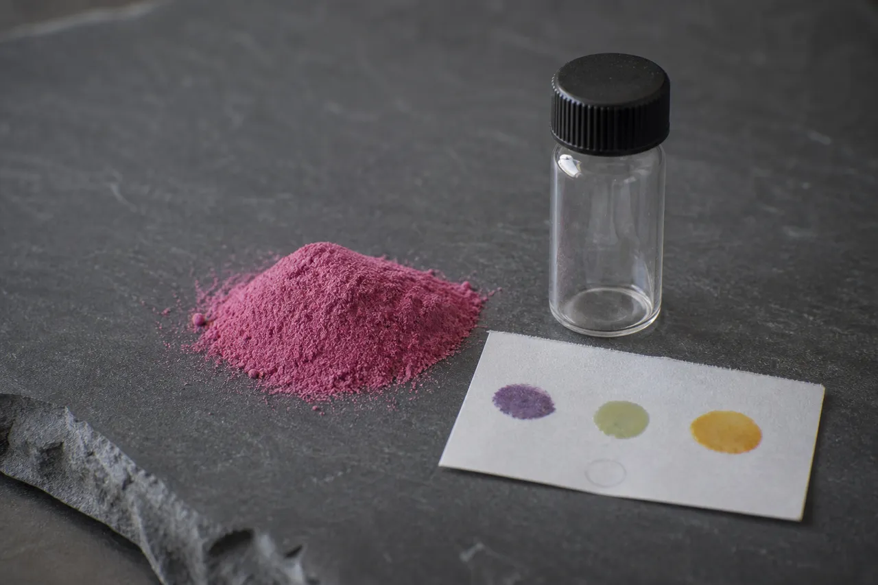 Editorial still life of a small mound of fine pink powder on a dark slate surface beside a clear glass laboratory vial and a paper reagent test card showing purple, olive and orange droplet reactions